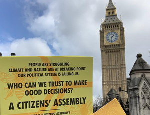 A yelow citizens assembly flyr is held up in front of Big Ben