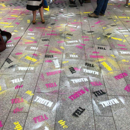 An inage of a pavement, with 'Tell' 'The' and 'Truth' stencilled all over it in white, pink, yellow and black spray paint