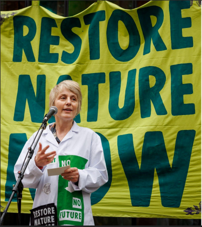A white female scientist in a lab coat adorned with patched stands at a microphone, in front of a yellow banner reading 'Restore Nature Now'