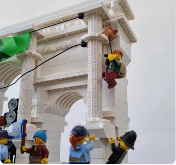 A lego swampy climbs a lego Marble Arch to hand a large XR banner. Lego police are standing at the bottom with their arms up in the air'